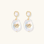 Gold-plated drop earrings with iridescent baroque pearls and crystal flower accents on cream background